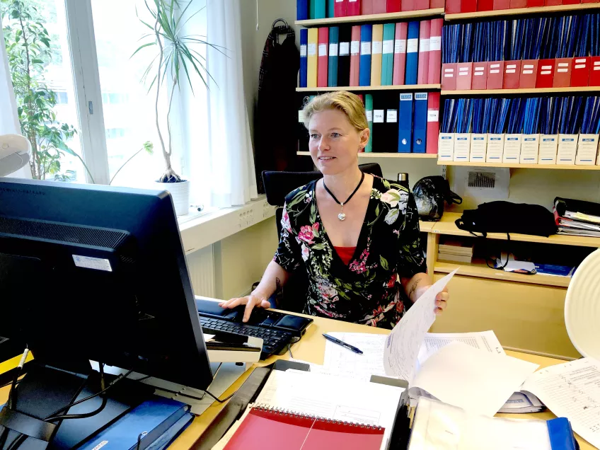Maria Månsson-Martinez vid datorn på sitt kontor. Bakom henne är bokhyllor med pärmar och på skrivbordet ligger pappersformulär. Foto.