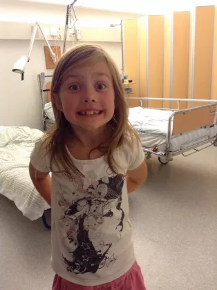 En flicka i ett sjukhusrum som tittar in i kameran och ser glad ut. Foto.