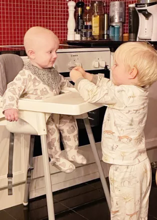 En pojke i pyjamas står vid sin lillebror som sitter i en barnstol och ger honom kex. Foto.
